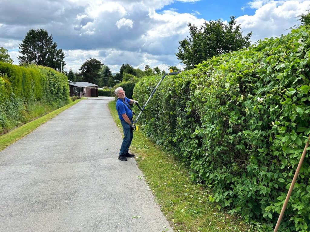 Ein Mann schneidet mit einer langen Heckenschere eine hohe grüne Hecke entlang eines Weges auf dem Arcanara-Land