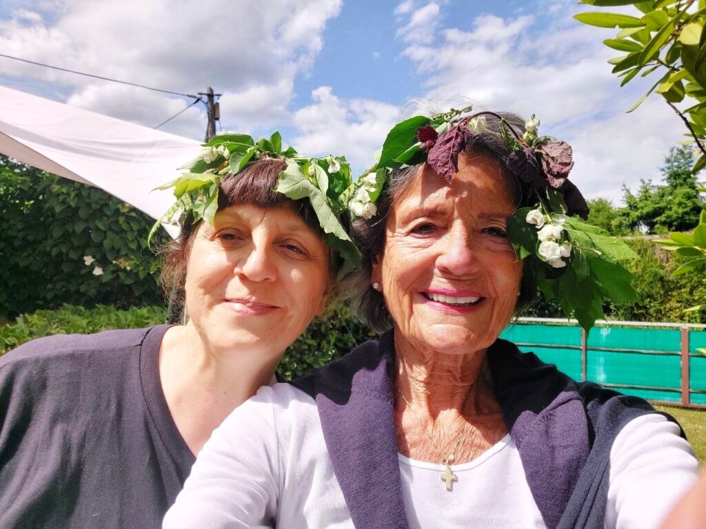 Zwei Frauen mit selbstgebundenen Kränzen aus Laub und weißen Blüten im Haar, lächelnd vor einer grünen Hecke unter blauem Sommerhimmel