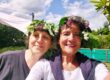 Zwei Frauen mit selbstgebundenen Kränzen aus Laub und weißen Blüten im Haar, lächelnd vor einer grünen Hecke unter blauem Sommerhimmel