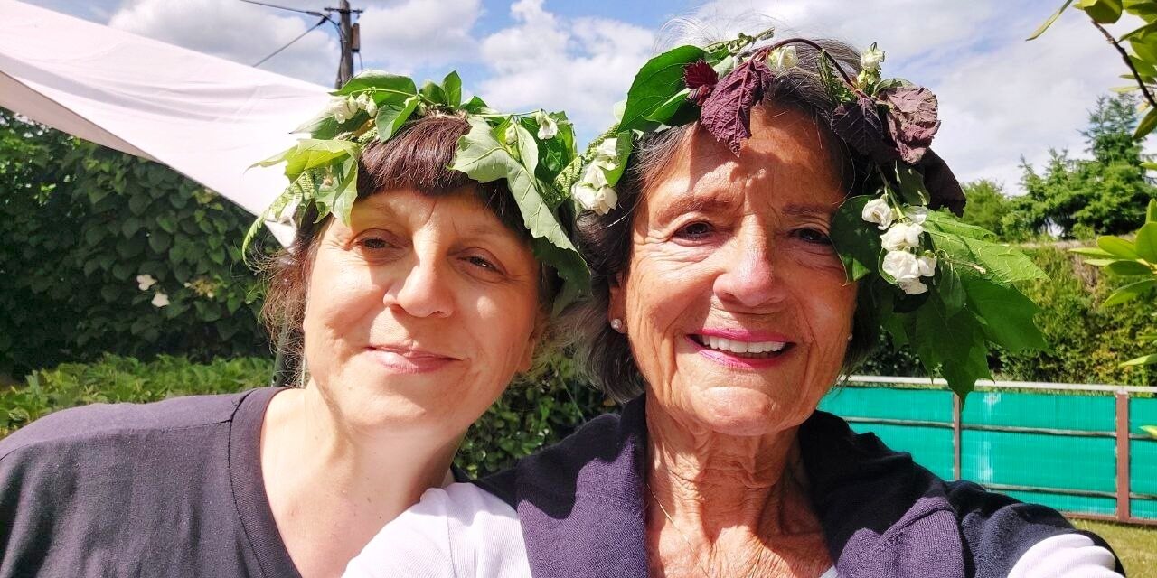 Zwei Frauen mit selbstgebundenen Kränzen aus Laub und weißen Blüten im Haar, lächelnd vor einer grünen Hecke unter blauem Sommerhimmel
