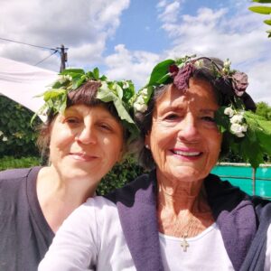Zwei Frauen mit selbstgebundenen Kränzen aus Laub und weißen Blüten im Haar, lächelnd vor einer grünen Hecke unter blauem Sommerhimmel