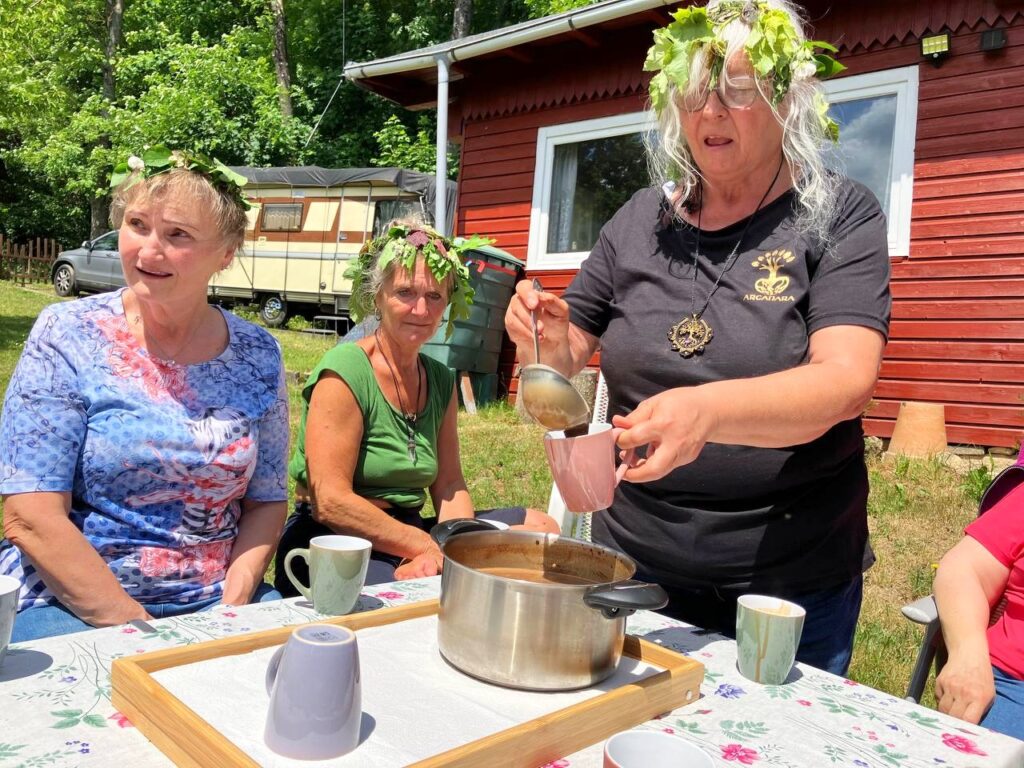 Eine Frau mit Laubkranz schöpft mit einer Kelle Kakao aus einem großen Topf, um sie herum sitzen Teilnehmerinnen an einem gedeckten Tisch vor einem roten Holzhaus
