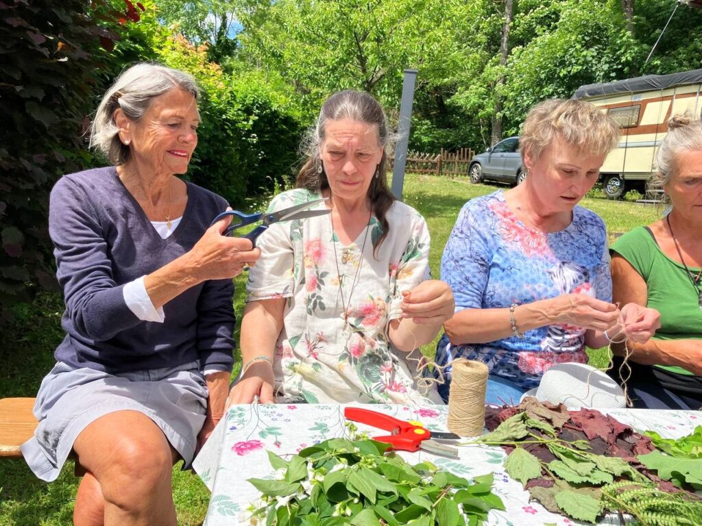 Vier Frauen sitzen um einen Gartentisch und binden gemeinsam Kränze aus Blättern und Blüten, auf dem Tisch liegen Laub und eine rote Gartenschere