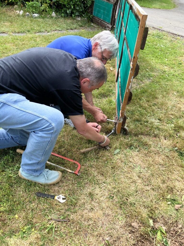Zwei Männer knien im Gras und schrauben gemeinsam an einem Rad, daneben liegt ein roter Wagenheber