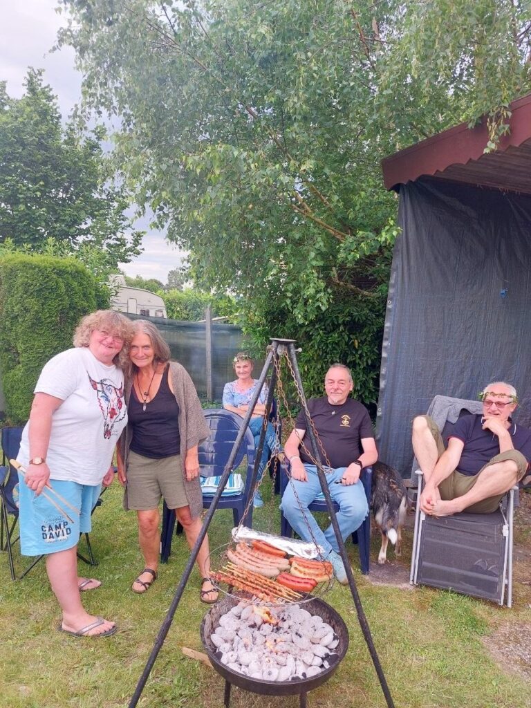 Über der Glut eines Schwenkgrills liegen Würstchen, dahinter stehen und sitzen mehrere Menschen in entspannter Runde unter Bäumen