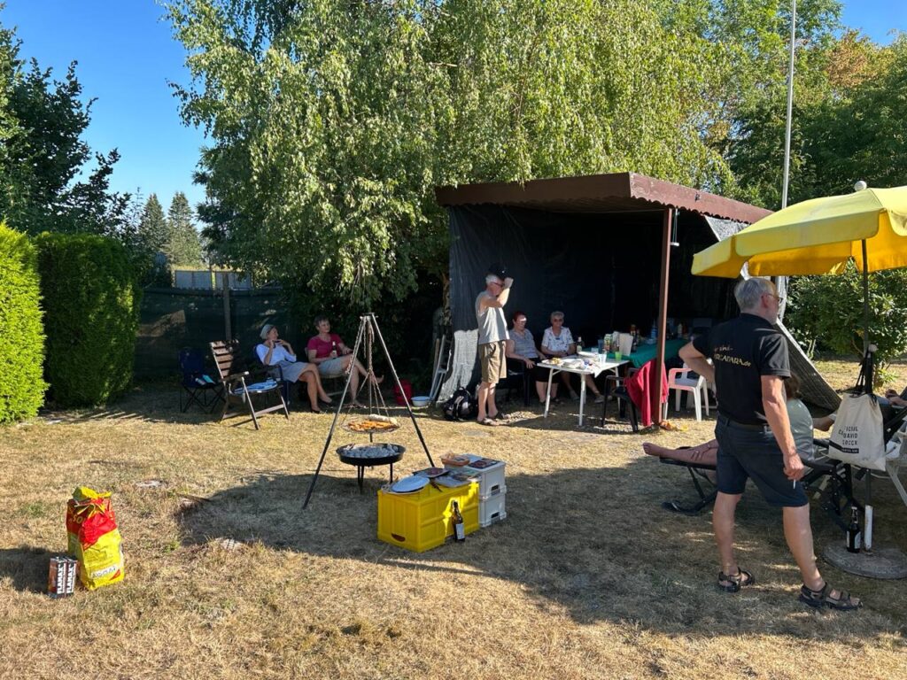 Sonniger Garten mit Schwenkgrill, Zeltdach und gelbem Sonnenschirm, im Hintergrund die Runde am Tisch