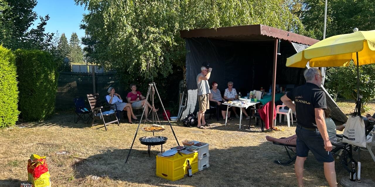 August-Treffen 2026 – im Garten Sonniger Garten mit Schwenkgrill, Zeltdach und gelbem Sonnenschirm, im Hintergrund die Runde am Tisch