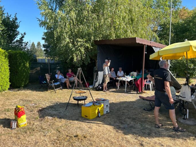 August-Treffen 2026 – im Garten Sonniger Garten mit Schwenkgrill, Zeltdach und gelbem Sonnenschirm, im Hintergrund die Runde am Tisch